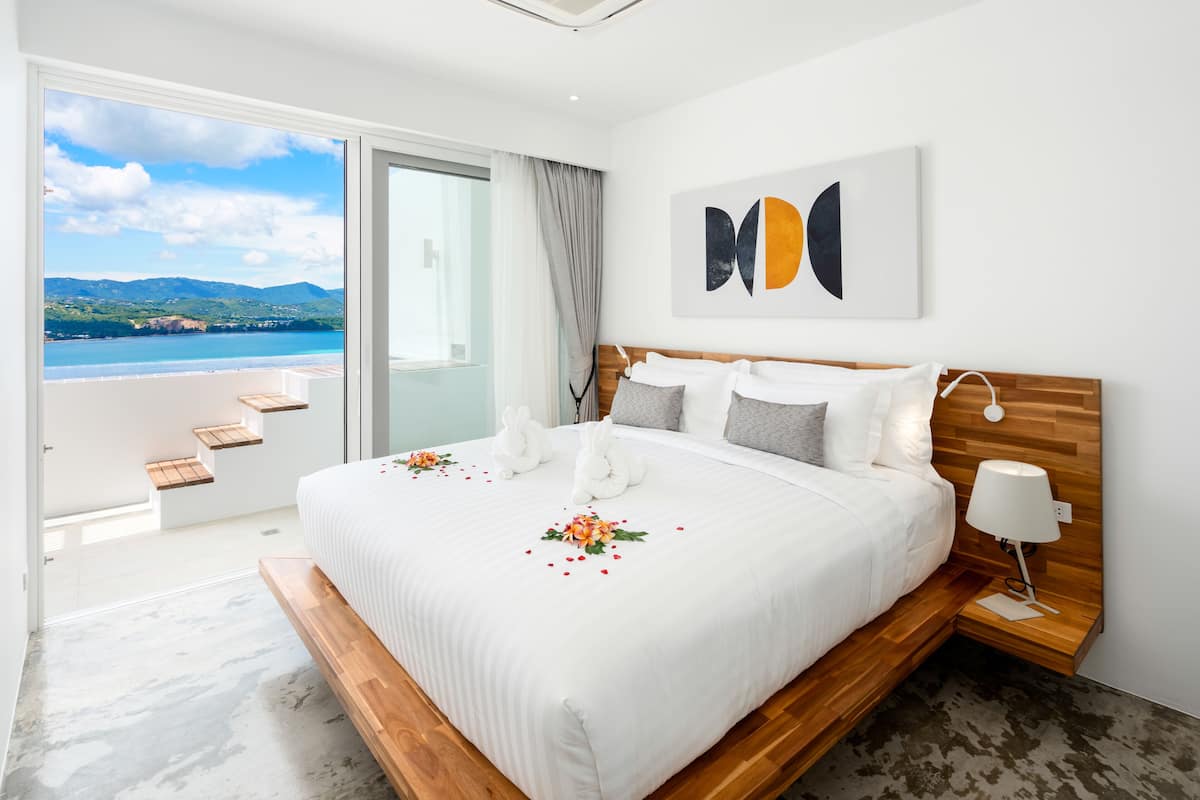 A hotel room with a large bed, a wooden headboard, a bedside table with a lamp, and a view of the sea and mountains through a sliding glass door.