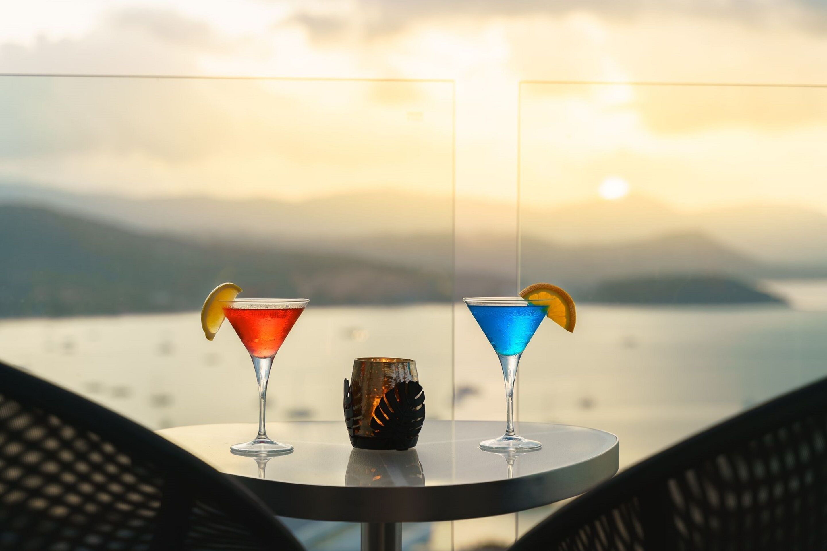Two cocktails on a table with a view of a body of water and mountains in the background.
