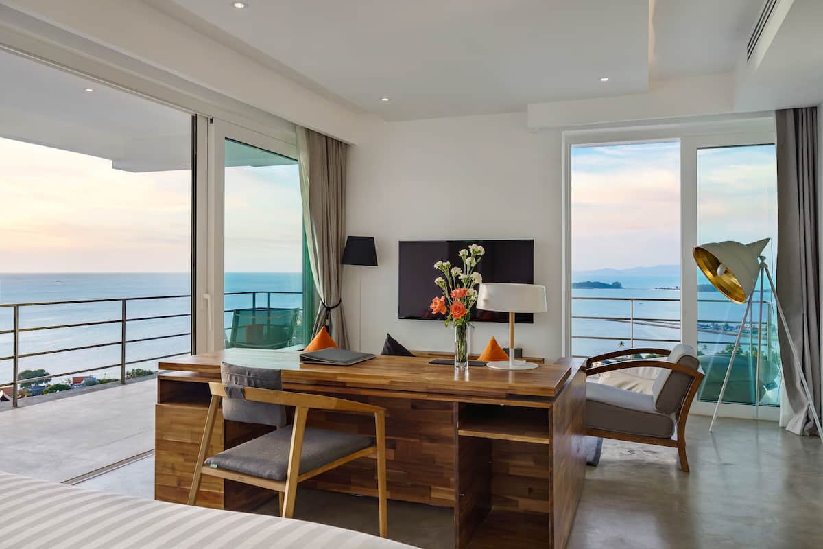 A modern hotel room with a wooden desk, two chairs, a flat-screen TV, and a large window offering a view of the sea.