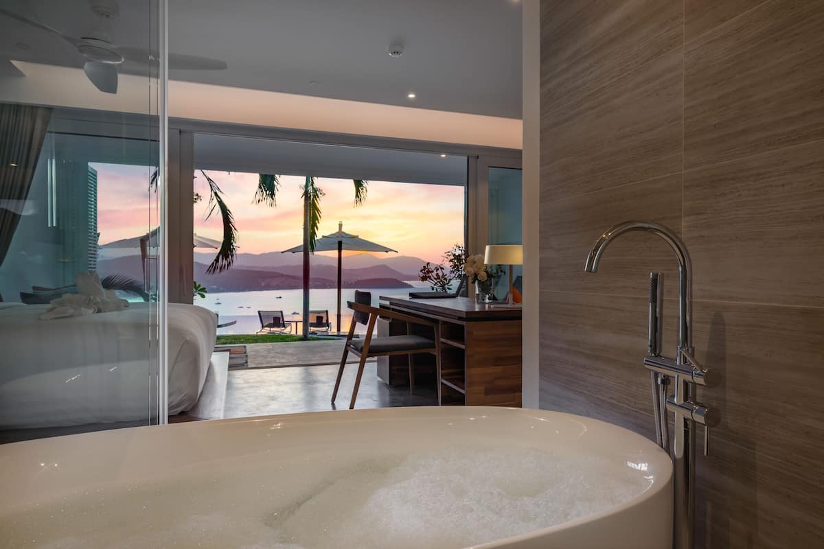 A modern bathroom with a large bathtub, a shower, and a view of a sunset over mountains.