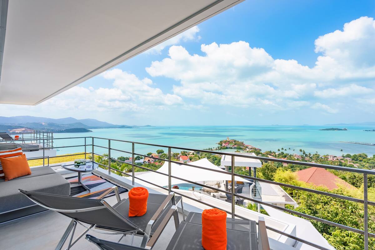 A balcony with a view of a turquoise sea and distant islands, furnished with lounge chairs and orange cushions.