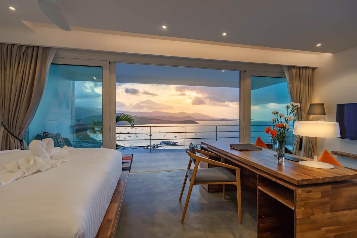 A hotel room with a large bed, a wooden desk, and a view of the sea and mountains.