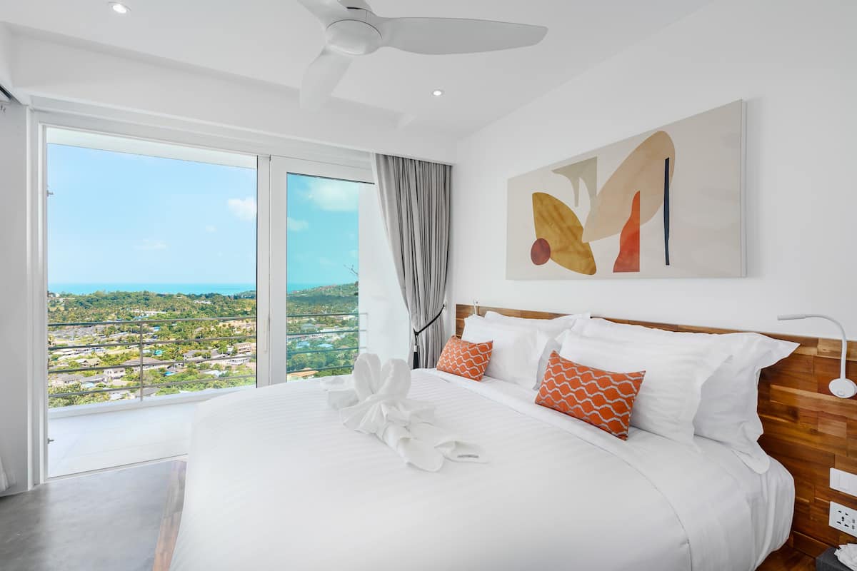 A modern hotel room with a large bed, a view of the city, and a ceiling fan.