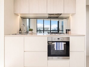 A modern kitchen with a built-in oven, stainless steel appliances, and a window overlooking a cityscape. - Kula Docklands (Docklands)