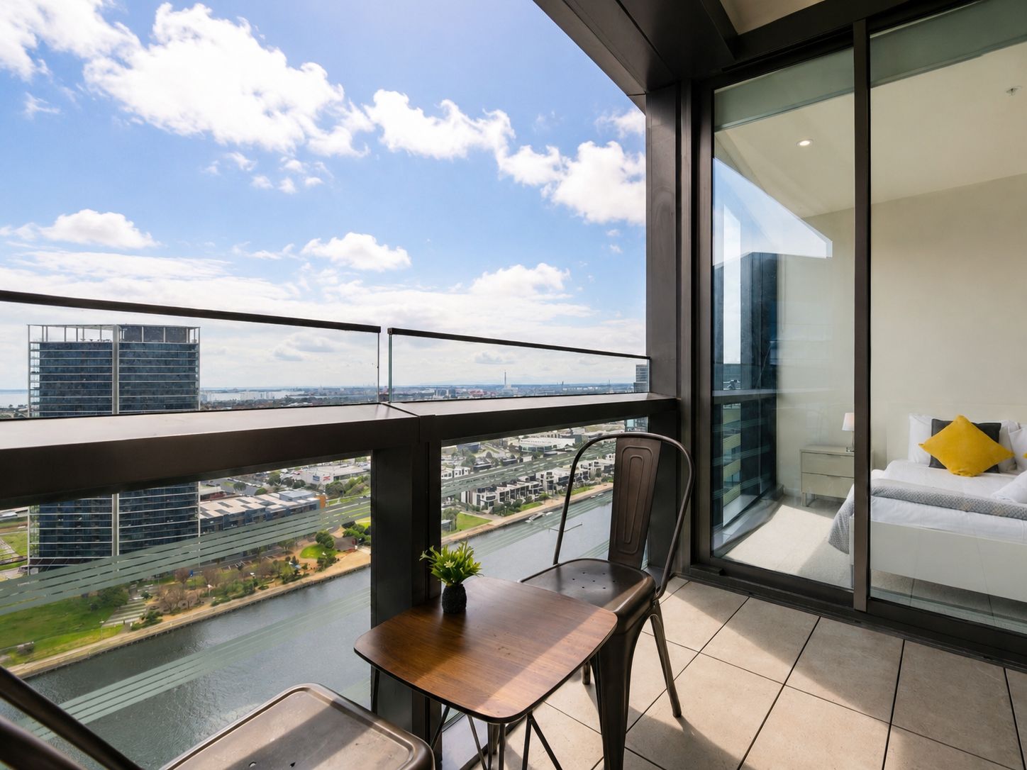 A balcony with a view of a river and cityscape, a dining area with a table and chairs, and a bedroom with a bed and a window.