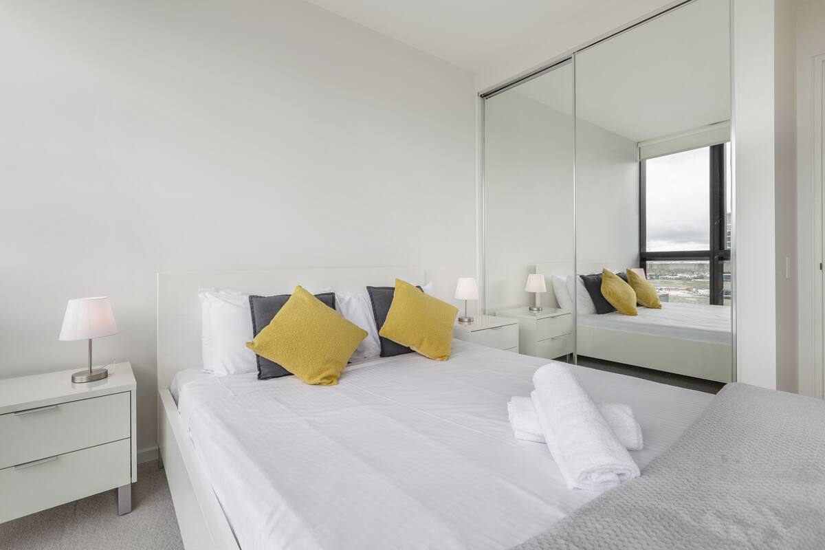 Ein modernes Hotelzimmer mit einem großen Bett, weißer Bettwäsche, einem Nachttisch mit Lampe sowie einem weiteren Beistelltisch und einem großen Spiegel, der den Raum reflektiert.