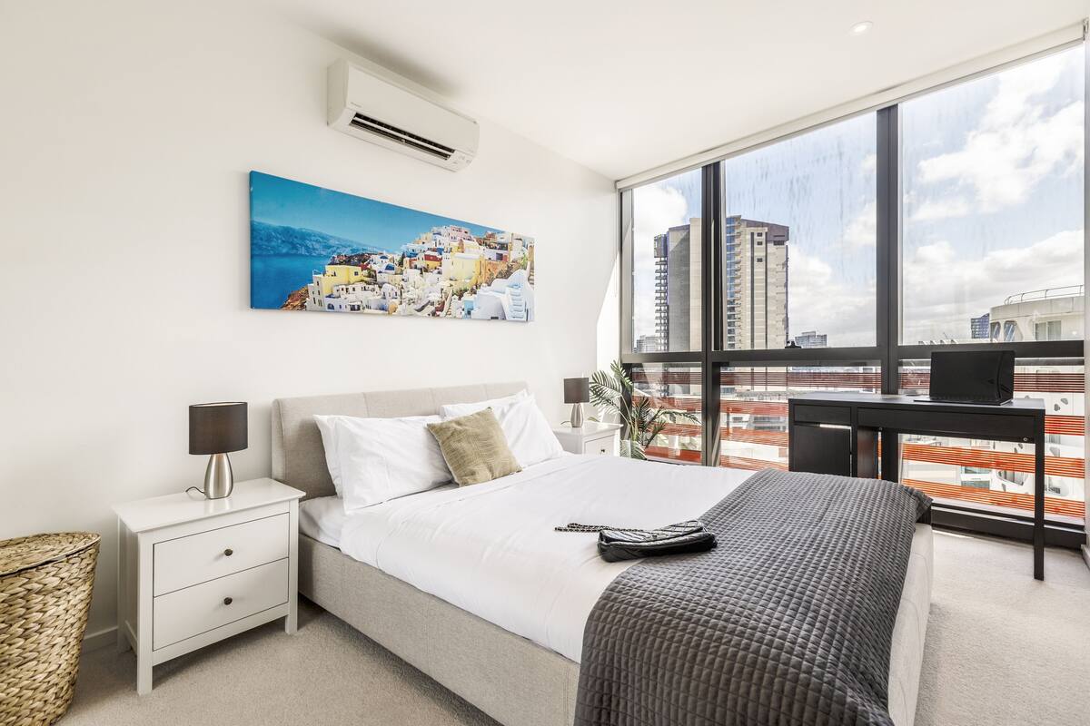 Ein modernes Hotelzimmer mit einem großen Bett, einem Schreibtisch und Blick auf die Stadt durch große Fenster.