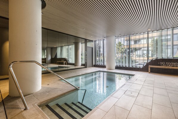 Une piscine intérieure avec des parois en verre transparent, un panneau indiquant qu’il est interdit de plonger et un sol carrelé.