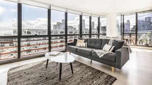 Una sala de estar moderna con un sofá gris, una mesa de centro blanca y un tapete estampado. Amplias ventanas ofrecen vistas a un paisaje urbano y a un tren.