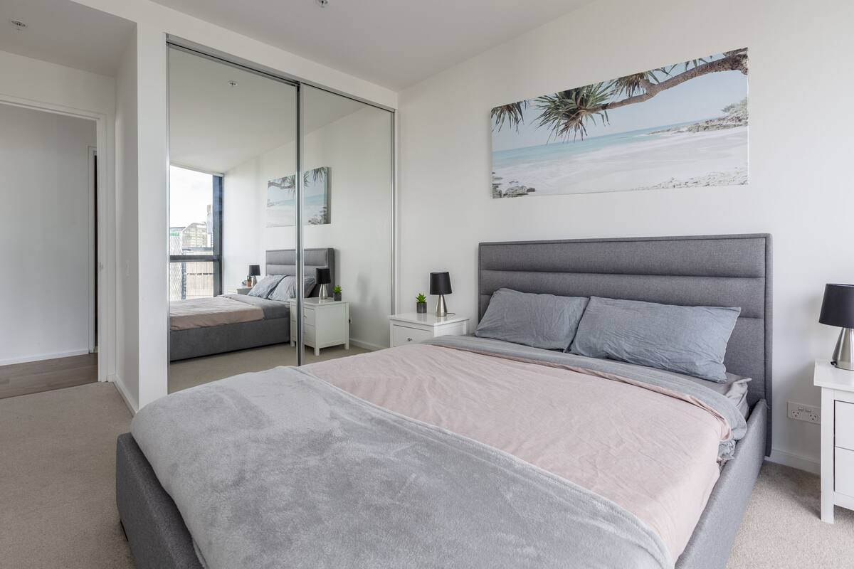 Ein modernes Schlafzimmer mit einem großen Bett, einer Schiebetür zu einem Balkon, einem Nachttisch mit einer Lampe und Wandbildern mit Strandmotiv.