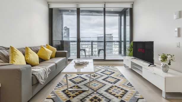 Una sala de estar moderna con sofá, mesa de centro, televisor y vistas a la ciudad a través de grandes ventanales.