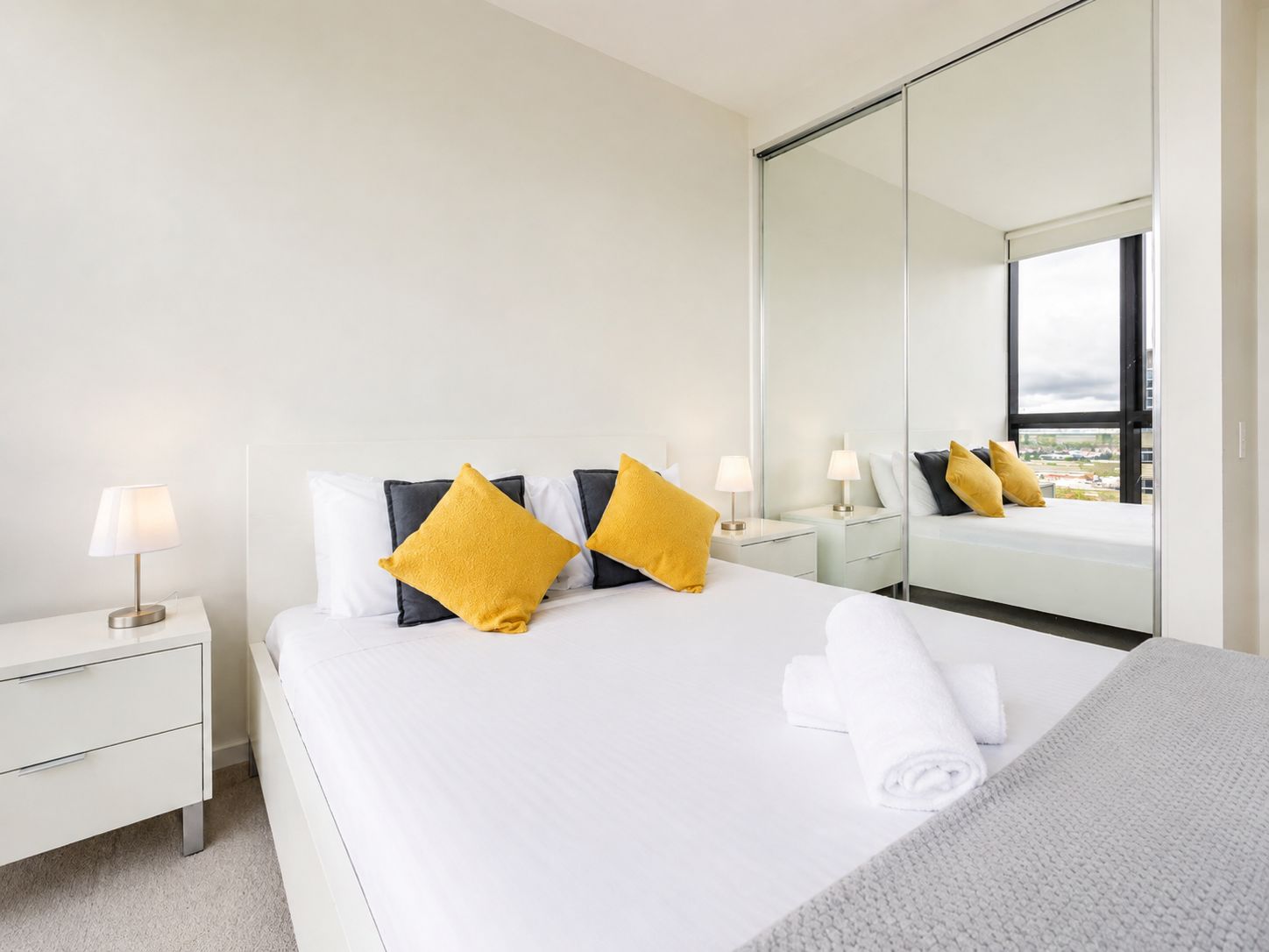 A hotel room with a bed, a nightstand, a lamp, a mirror, and a view of the city.