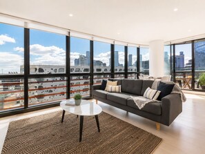 A modern living room with a large window, a gray sofa, a small round table, and a potted plant. - Kula Docklands (Docklands)