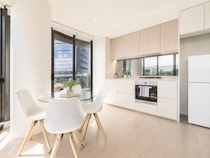 Une cuisine moderne avec un coin repas, comprenant une table en verre, des chaises blanches et une vue sur la ville grâce à de grandes fenêtres.