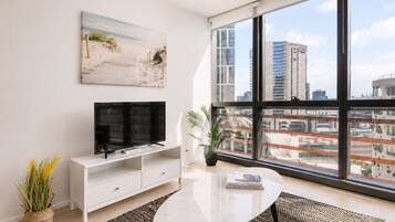 A modern living room with a large window, a flat-screen TV, a white entertainment unit, a round coffee table, and a beach-themed painting.