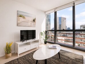 A modern living room with a large window, a flat-screen TV, a white entertainment unit, a round coffee table, and a beach-themed painting. - Kula Docklands (Docklands)