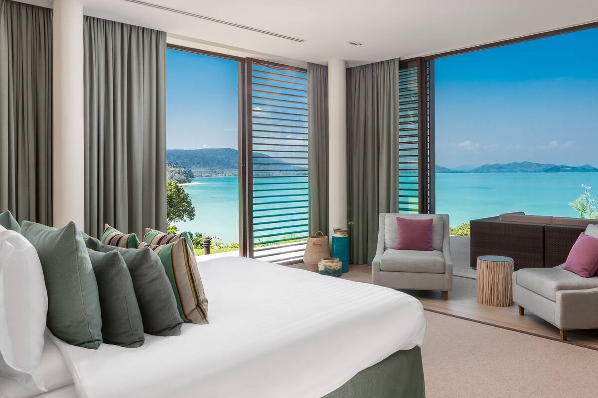 A modern hotel room with a large bed, two seating areas, and a view of a turquoise sea and distant mountains.