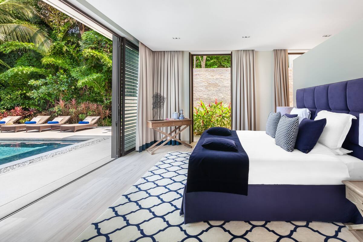 A modern hotel room with a large bed, a bedside table, a lamp, and a view of a swimming pool and greenery.