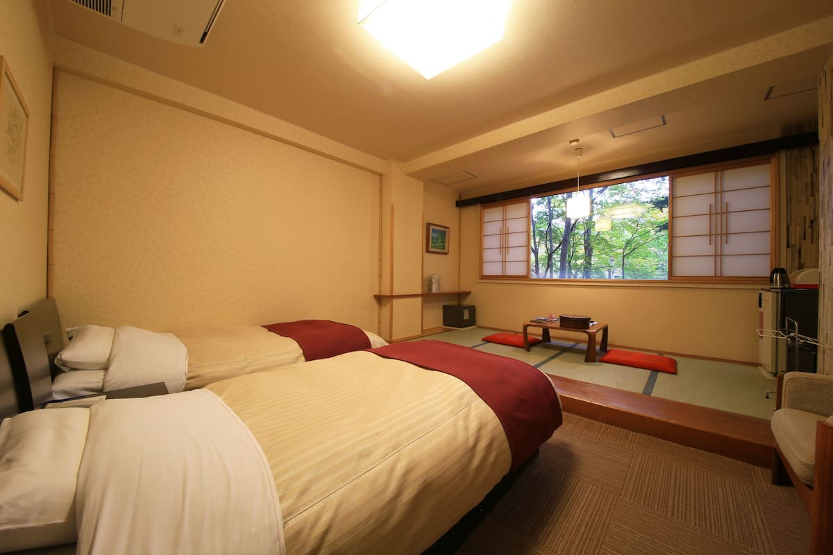 A hotel room with two beds, a small table, a chair, a window with a view of trees, and a sliding door.