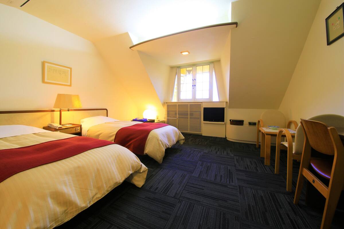 A hotel room with two beds, a desk, a chair, a television, and a window with curtains.