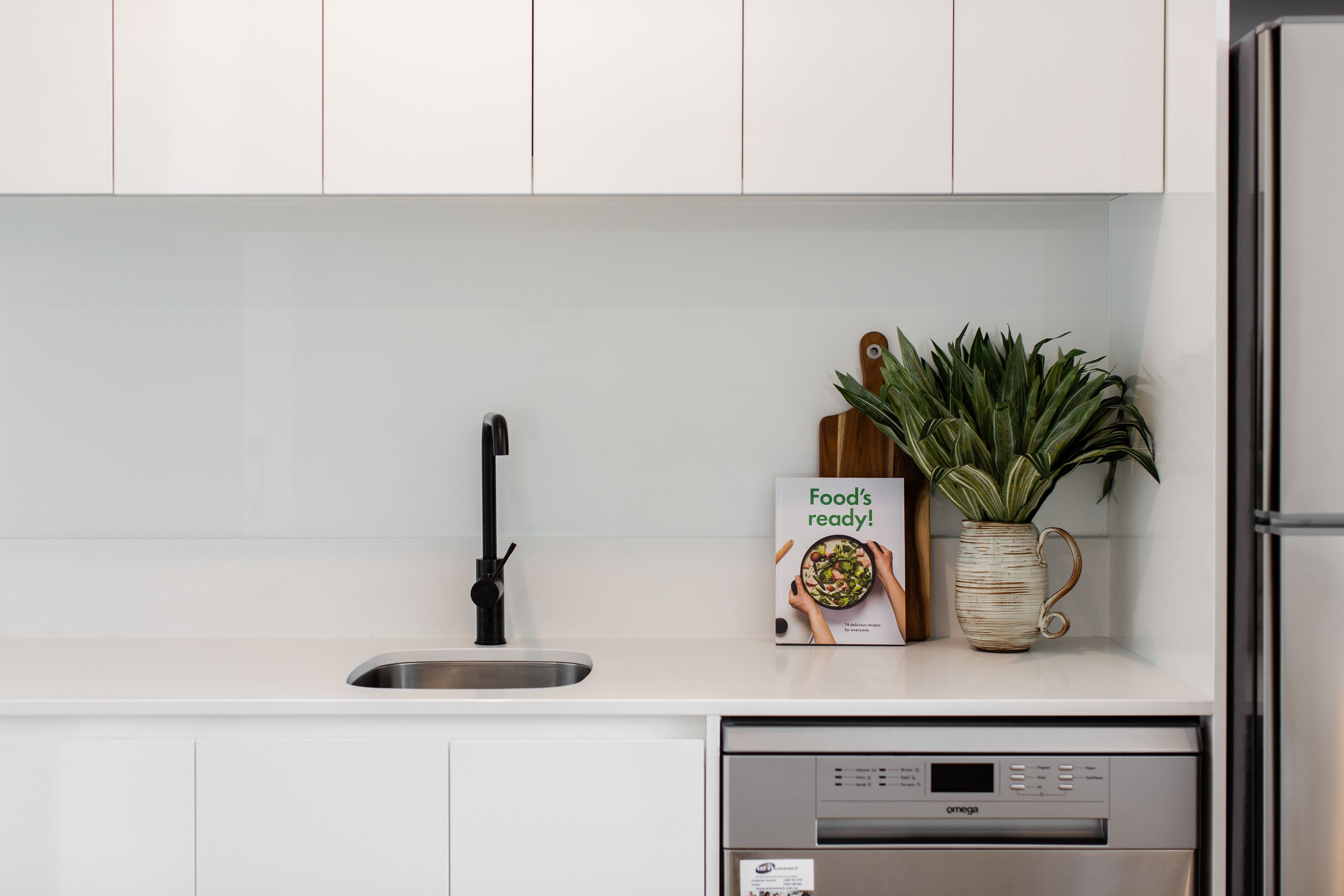 Una cocina moderna con armarios blancos, un lavamanos de acero inoxidable, lavavajillas y una planta en maceta.
