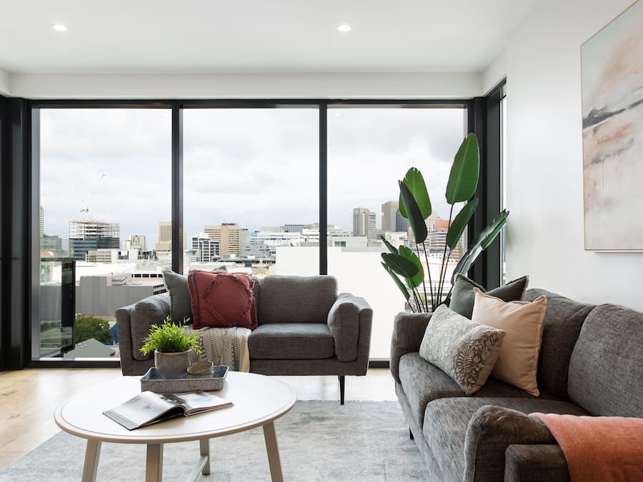 A modern living room with a large window, a grey sofa, a coffee table, and a potted plant.