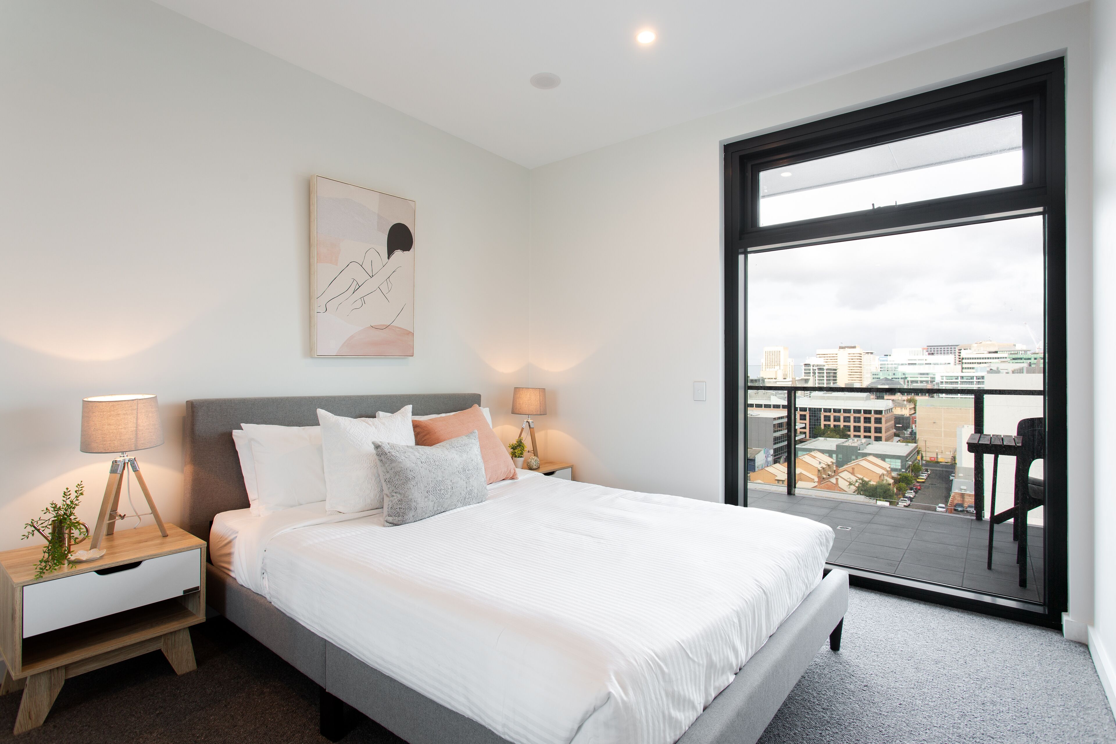 A modern bedroom with a large bed, two bedside tables with lamps, a city view through a large window, and a framed artwork on the wall.