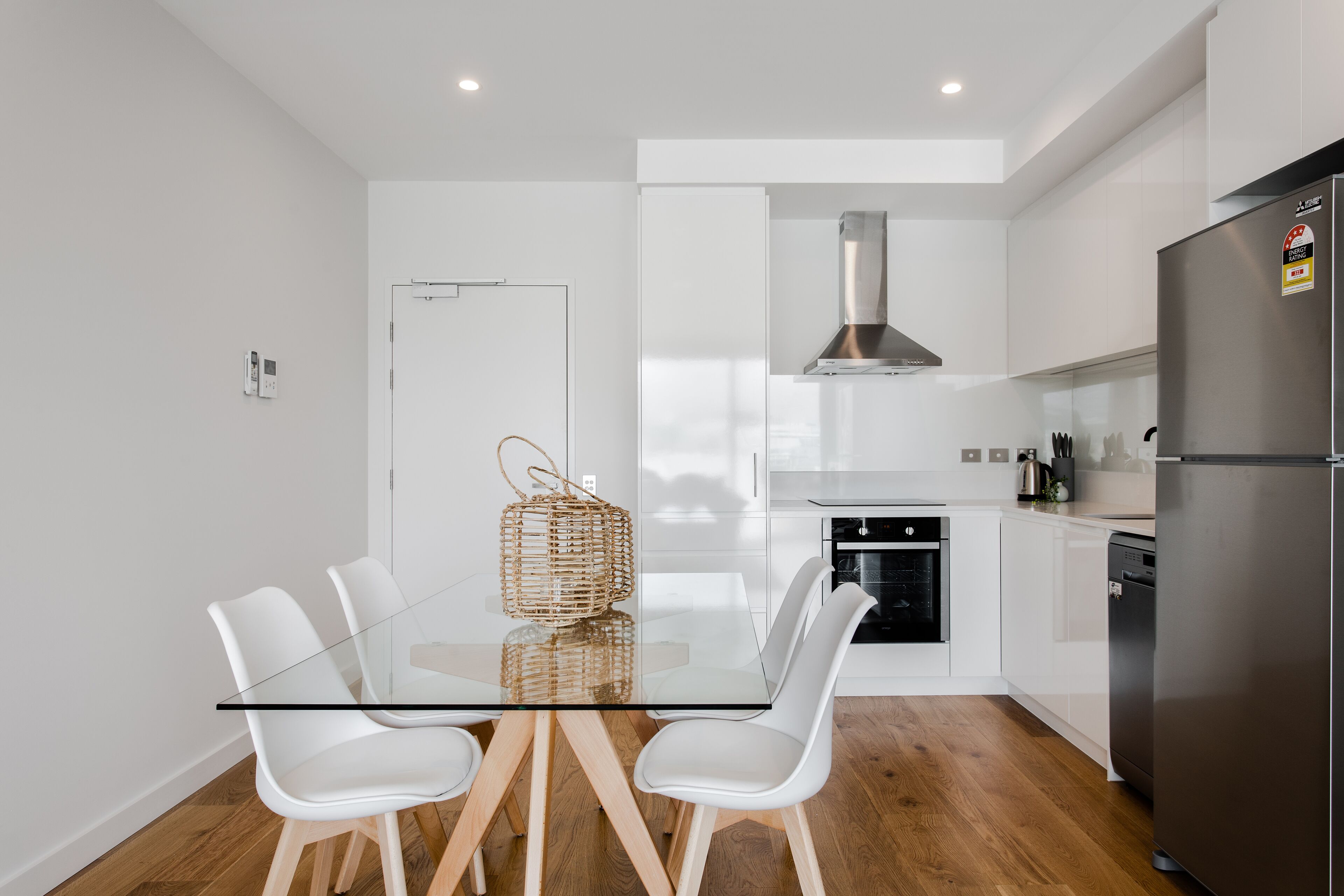 Una cocina moderna con una mesa de comedor de vidrio, sillas blancas, un refrigerador de acero inoxidable y un horno empotrado.