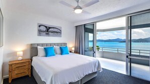 A modern hotel room with a large bed, a ceiling fan, a nightstand, and a view of the sea through large windows.