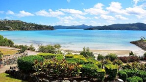 A coastal landscape with a clear blue sea, a sandy beach, and lush greenery in the foreground.
