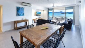 A modern living room with a wooden dining table, black chairs, a sofa, and a balcony with a view of the sea.