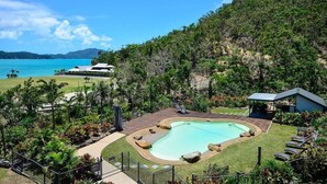 A swimming pool surrounded by a deck, with a view of the sea and hills in the background.