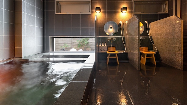 A modern spa with a large hot tub, wooden stools, and wall-mounted lights. - Super Hotel Kumamoto Yatsushiro (Yatsushiro)