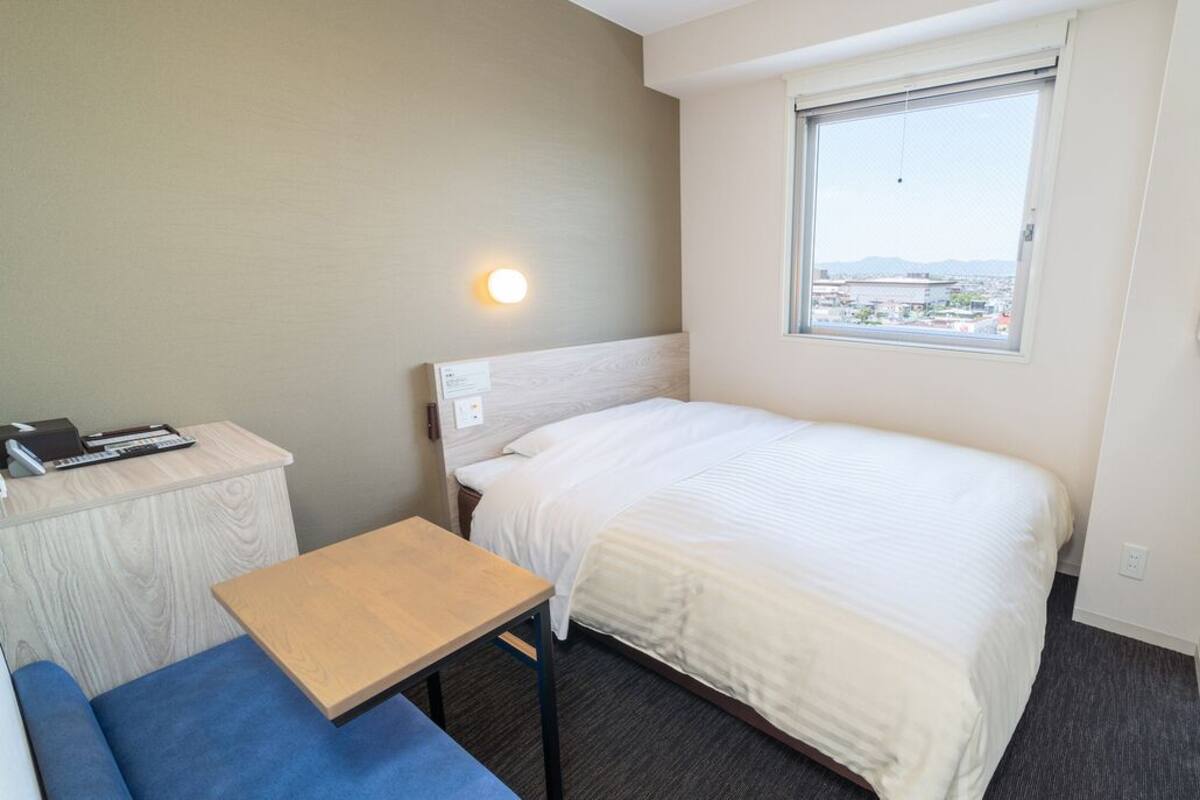 A small, clean room with a bed, a desk, a chair, and a window with a view.