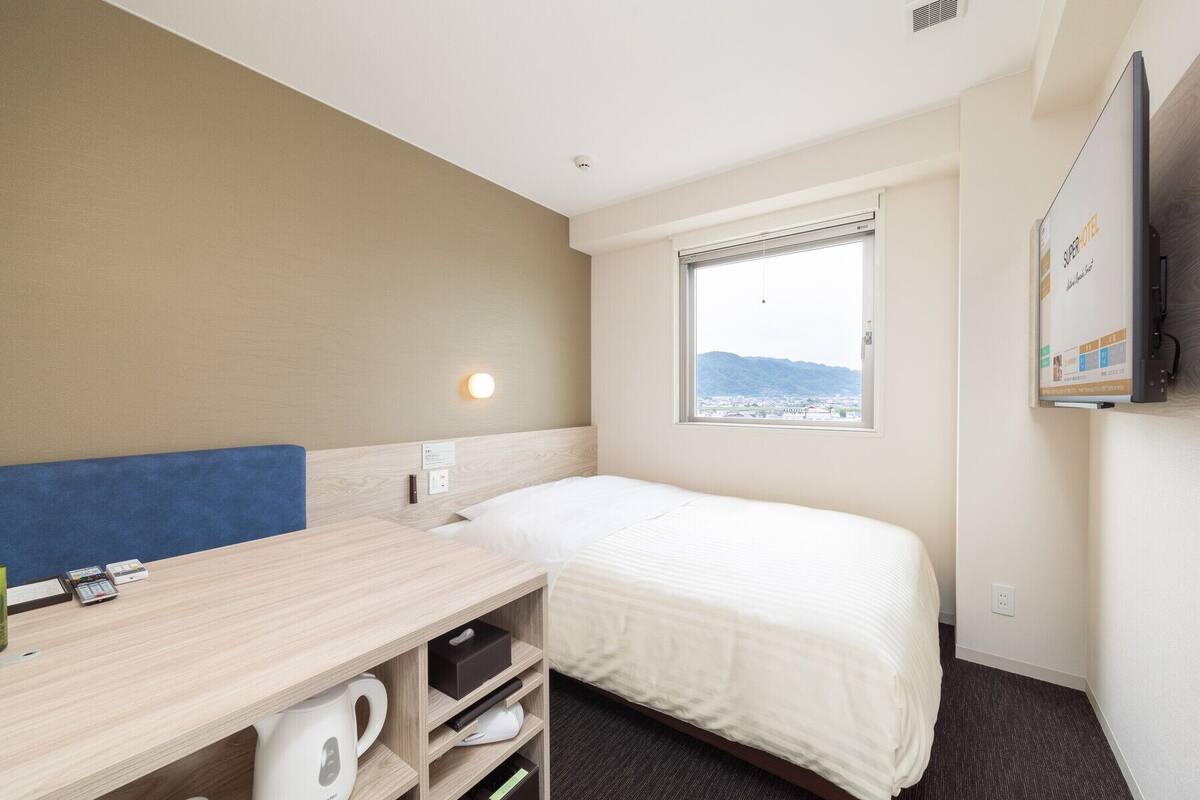 A hotel room with a bed, a desk with a kettle, a TV, and a window with a view of a city and mountains.