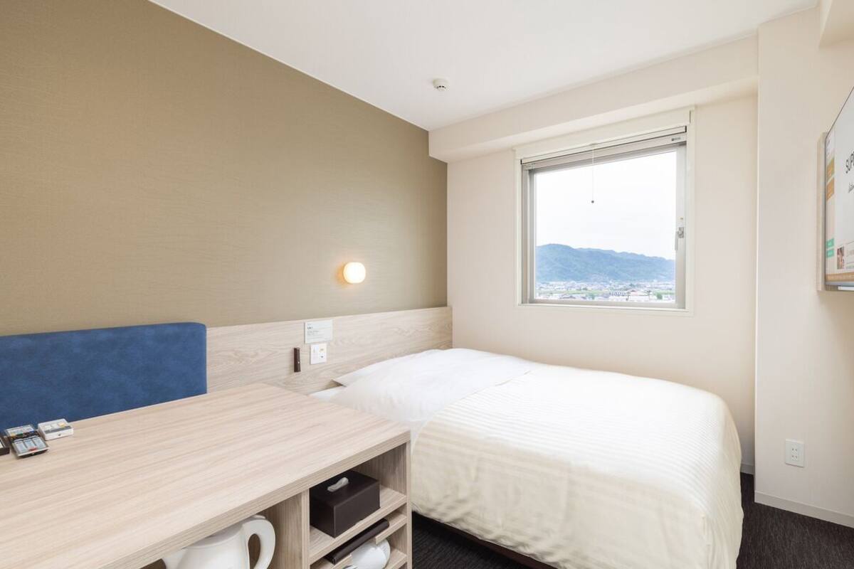 A hotel room with a single bed, a desk with a chair, a window with a view of the city, and a wall-mounted TV.
