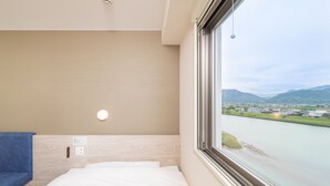 A hotel room with a bed, a blue towel, a wall-mounted light, and a window with a view of a river and mountains. - Super Hotel Kumamoto Yatsushiro (Yatsushiro)