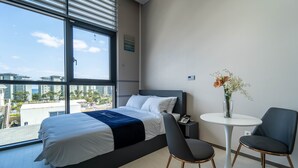 A modern hotel room with a large bed, two chairs, a small table, and a view of the city. - Hi Ocean Hotel Gyeongpo (Gangneung)