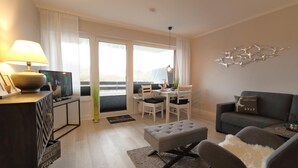 A modern living room with a sofa, ottoman, dining table, and chairs. There is a television on a stand, a decorative wall piece, and a view through the glass doors. - Apartment Widlak - Beach crossing K.Christiansen (Sylt)