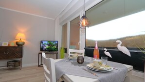 A dining area with a table set for two, a television in the background, and a window with a view of the outdoors. - Apartment Widlak - Beach crossing K.Christiansen (Sylt)