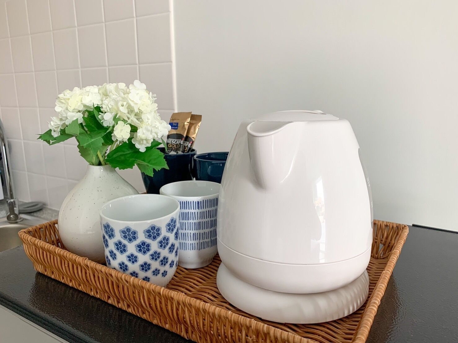 A kitchen counter with a white kettle, a white vase with white flowers, and two Nescafe Gold coffee cups on a woven tray.