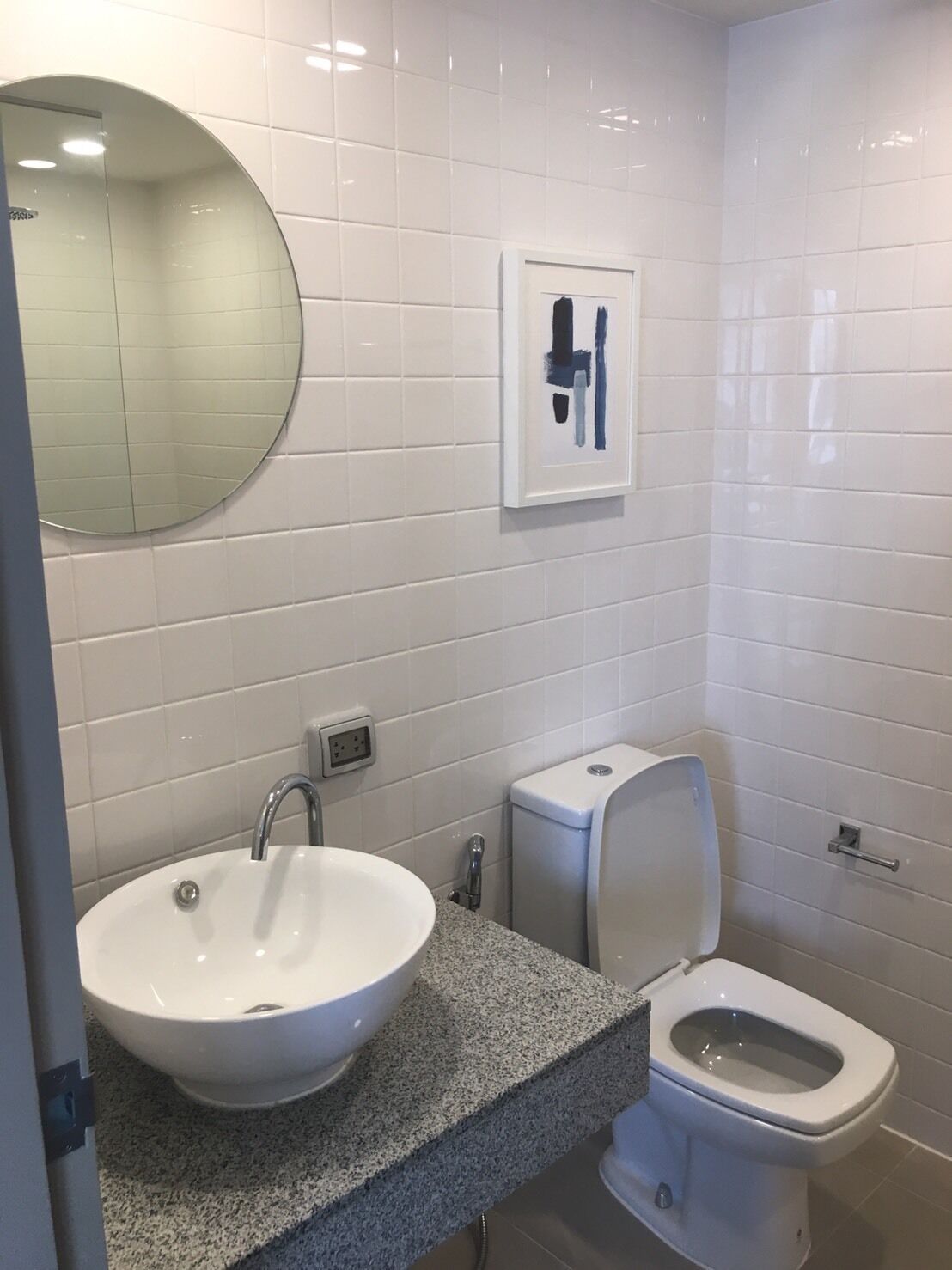 Un baño con un espejo redondo, un lavabo blanco, un inodoro y una obra de arte enmarcada en la pared.