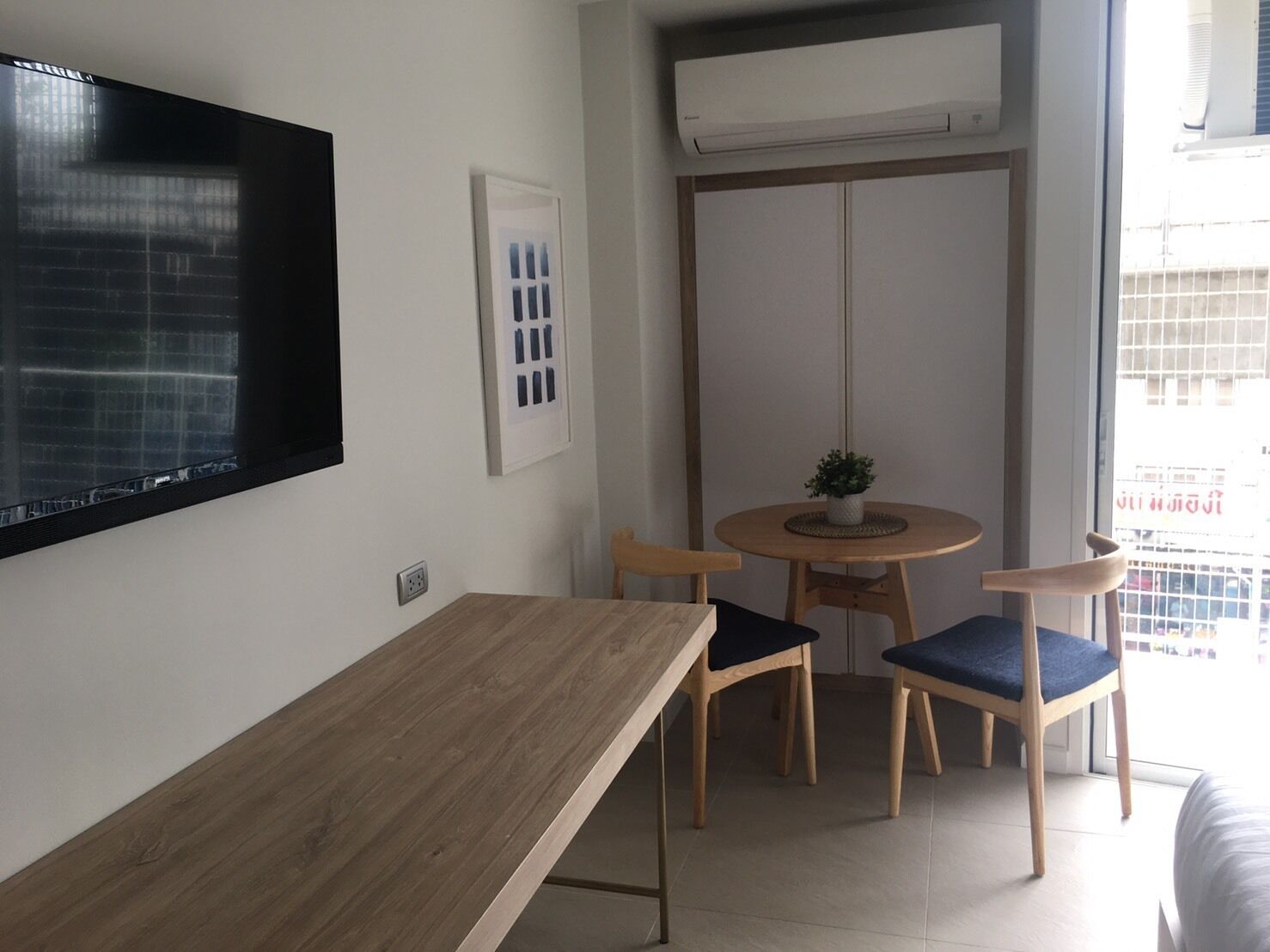 A small, modern room with a wooden table and two chairs, a mounted TV, an air conditioning unit, and a window with a view of buildings.