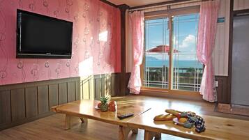 Een kamer met een houten tafel, een aan de muur bevestigde televisie, een raam met gordijnen en uitzicht op de zee.