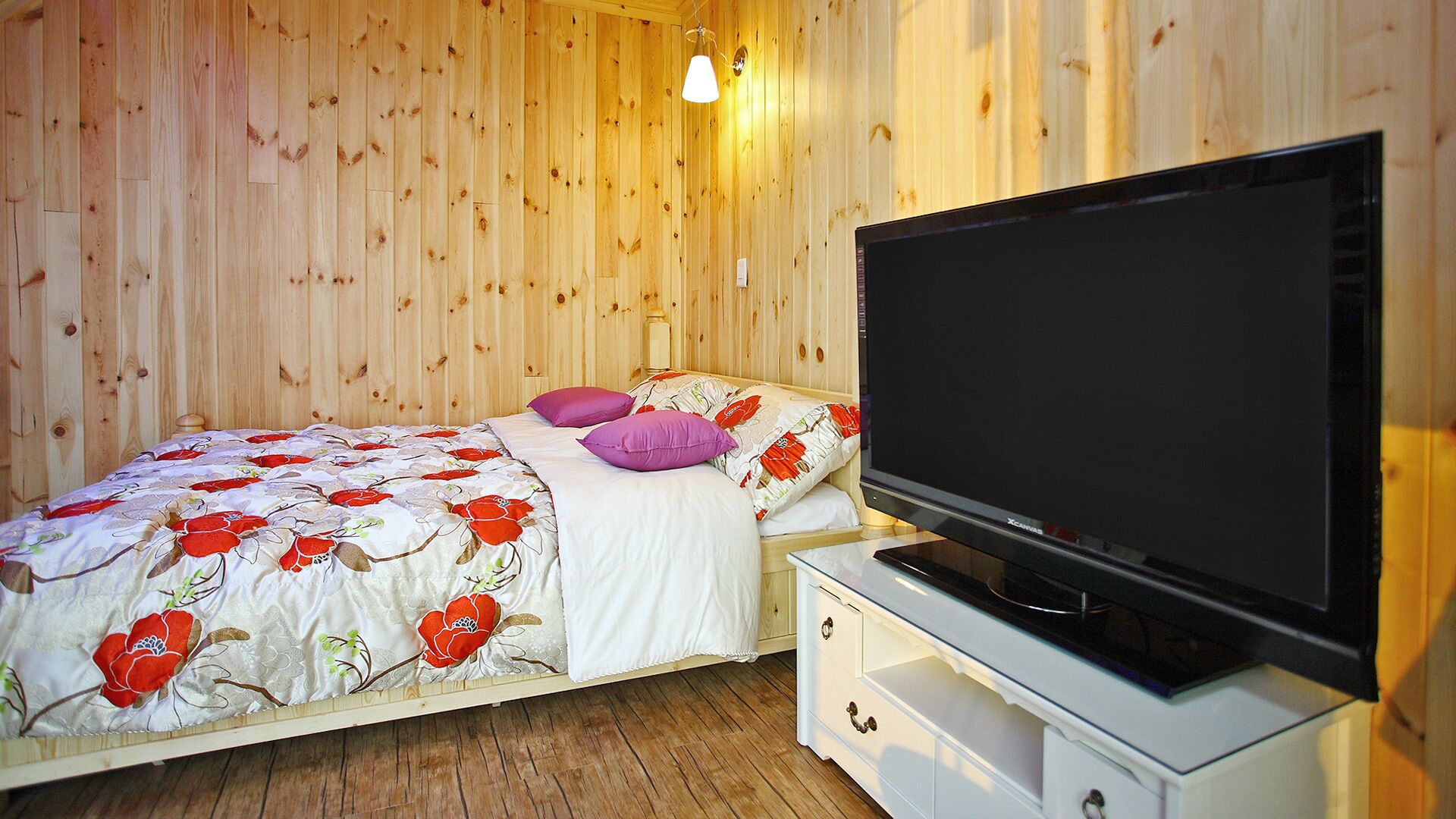 Un dormitorio con pared de madera, una cama con ropa de cama floral, dos almohadas, un televisor sobre un mueble bajo y un tocador pequeño blanco.