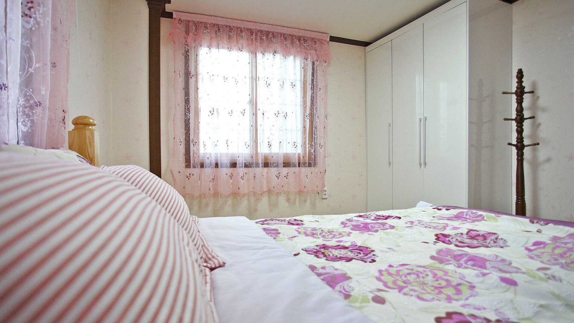 A bedroom with a bed, a window with sheer curtains, a wardrobe, and a coat stand.