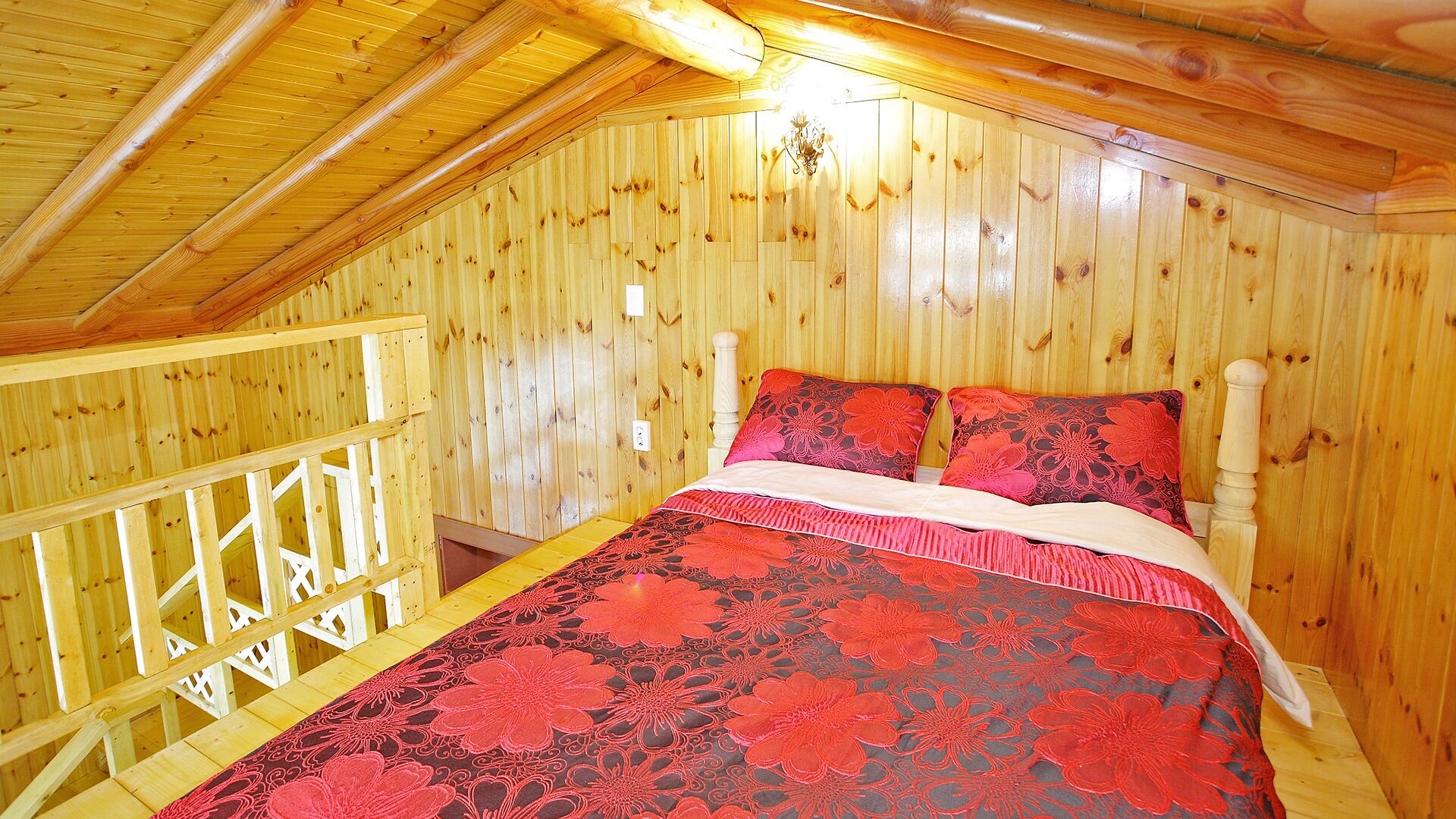A wooden cabin bedroom with a sloped ceiling, a wooden bed with floral bedding, and a balcony with a wooden railing.