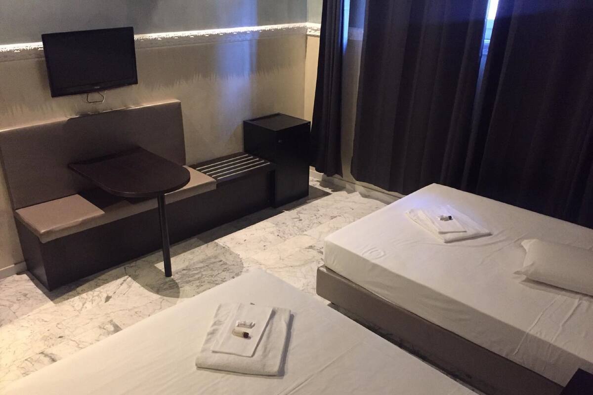 A hotel room with a sofa bed, a single bed, a TV mounted on the wall, and a small table.