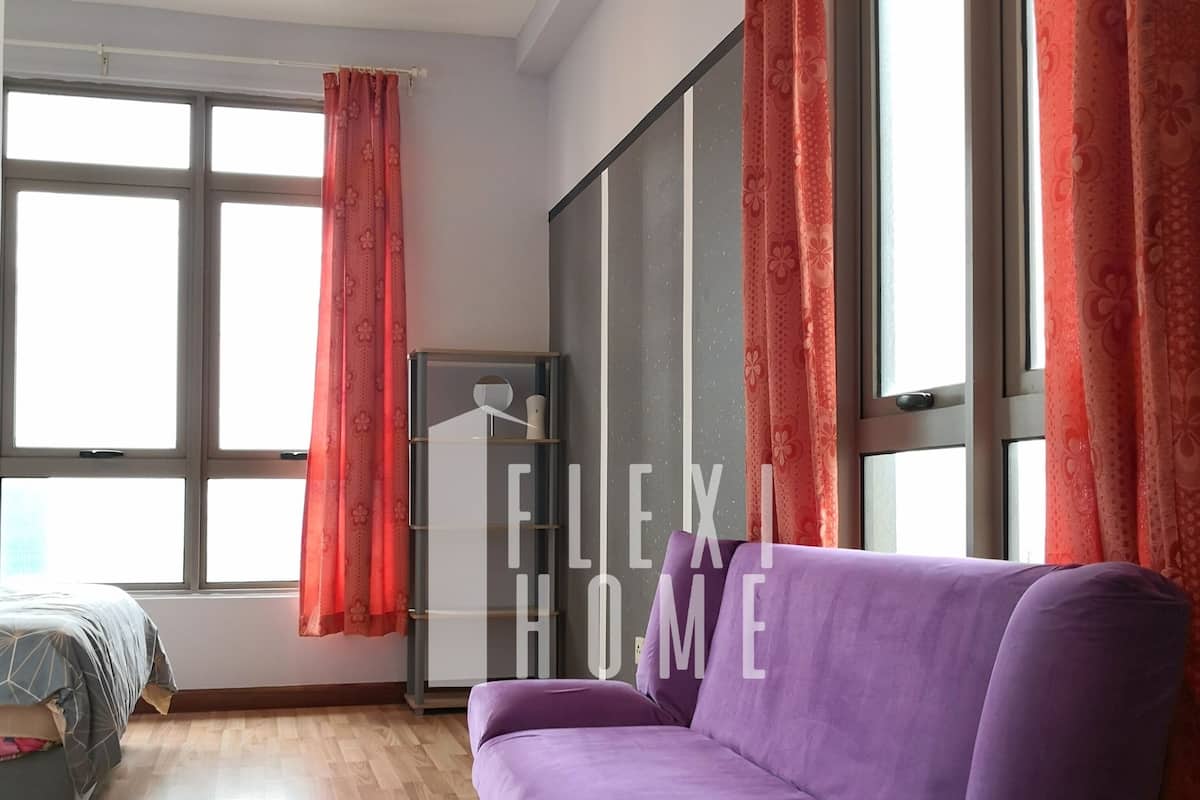A modern room with a purple sofa, large windows with red curtains, a bed with a grey bedspread, and a wooden floor.
