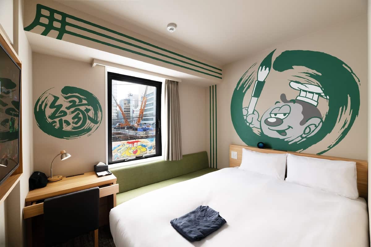 A hotel room with a bed, desk, and a window with a view of buildings and cranes.